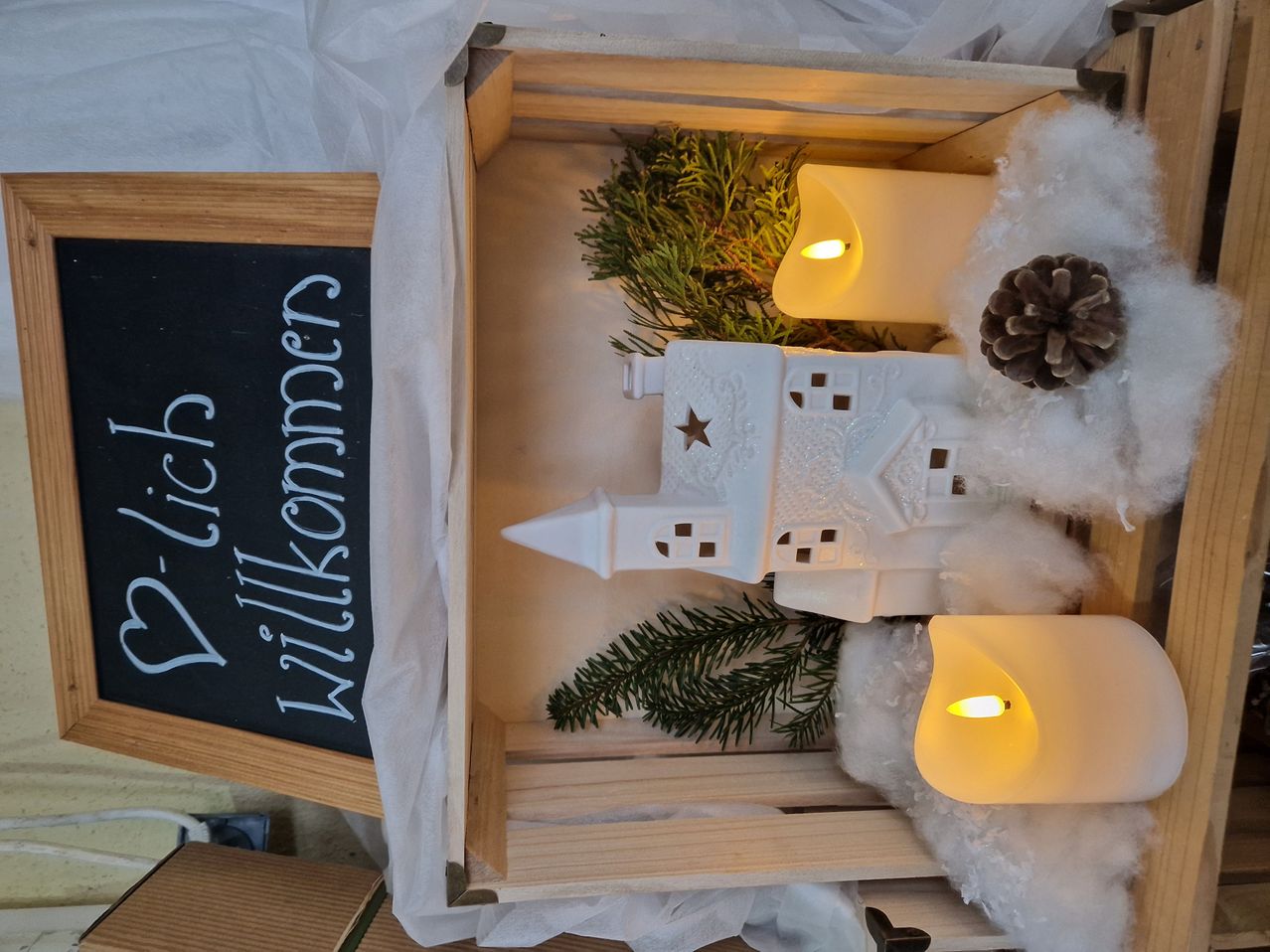 Eine Holzkiste mit einem weißen Keramikmodell einer Kirche, zwei brennenden Kerzen und einem Tafel mit 'Frohe Weihnachten' darauf. Es gibt Baumwolle und grüne Blätter um die Kirche herum.