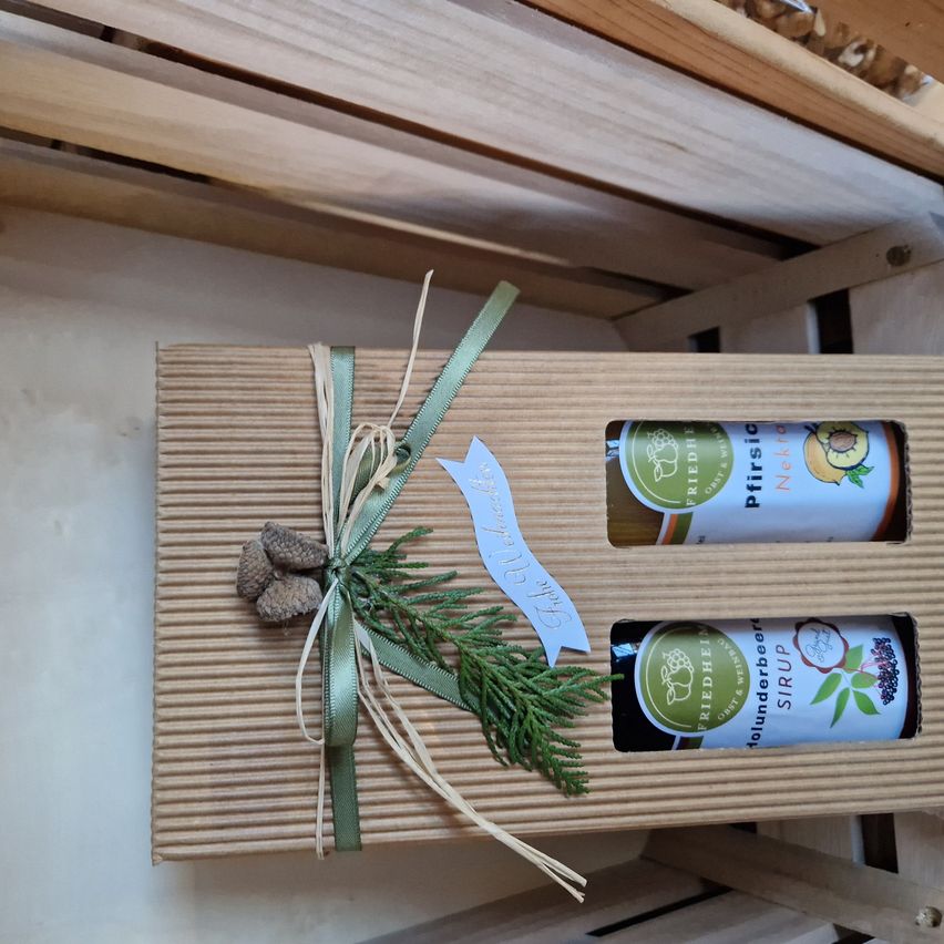 Ein Geschenkkarton mit zwei Flaschen Sirup, in einem grünen Band verpackt, geschmückt mit Tannenzapfen und grünen Blättern, auf einer Holzoberfläche präsentiert.