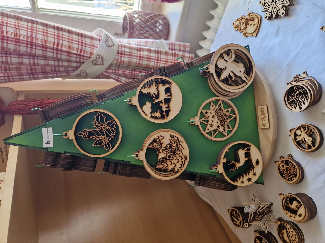 A green triangular board displays six wooden ornaments, each featuring a different design, including an angel, a star, and a reindeer. The ornaments are attached to the board with strings. In the background, there are other ornaments and a basket.