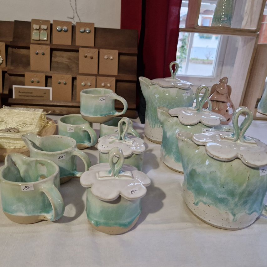 Eine Ausstellung von Keramik umfasst Teekannen, Tassen und Becher, alle in Grün- und Weißtönen bemalt. Sie sind auf einer weißen Tischdecke angeordnet, mit zusätzlichen dekorativen Elementen im Hintergrund.