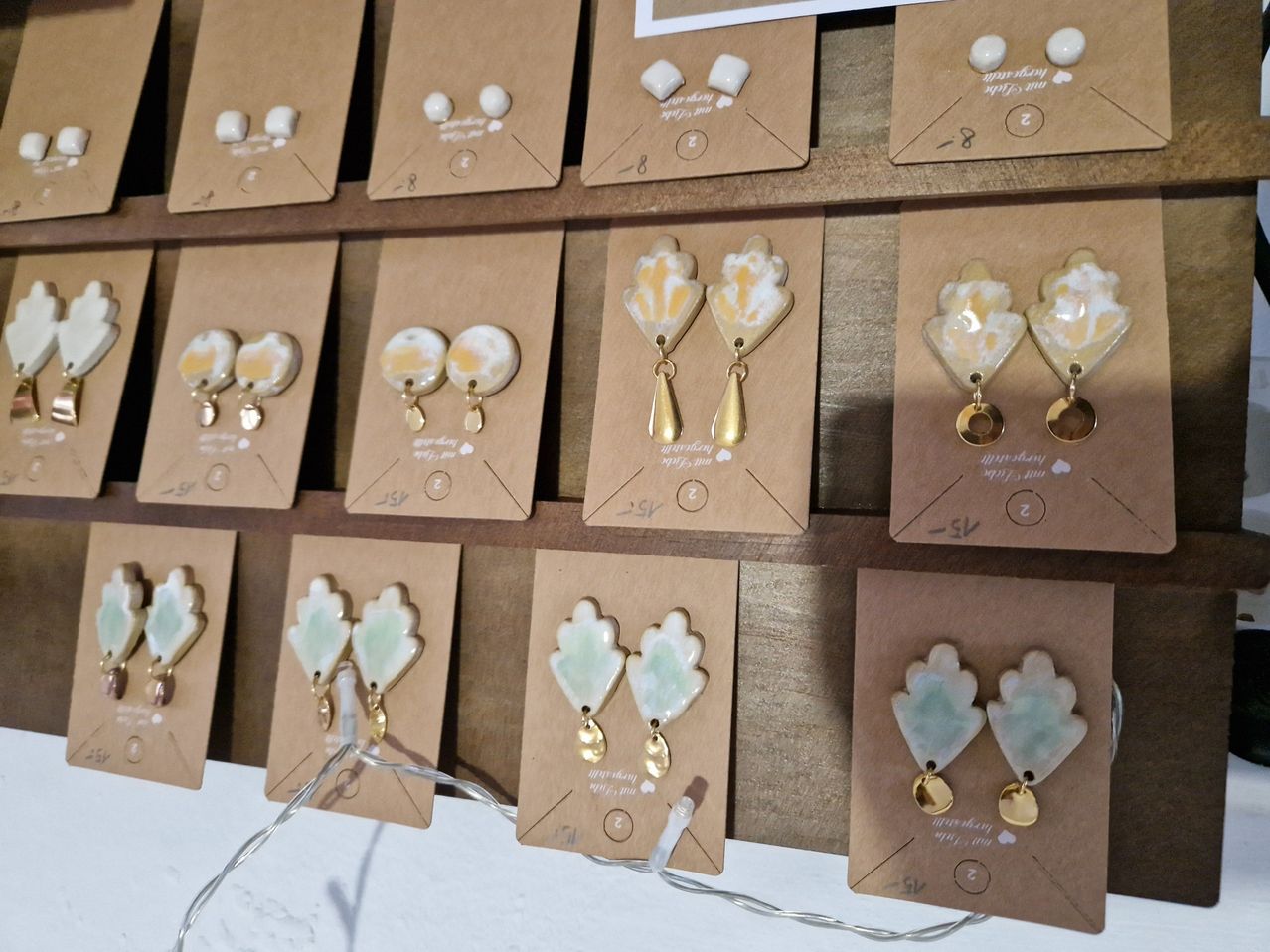 Ein Holzregal zeigt eine Auswahl an Keramik-Ohrringen. Jeder Ohrring ist einzigartig gestaltet, mit goldenen Akzenten und unterschiedlich gefärbten Steinen. Sie sind einzeln in braunen Umschlägen mit Etiketten verpackt.
