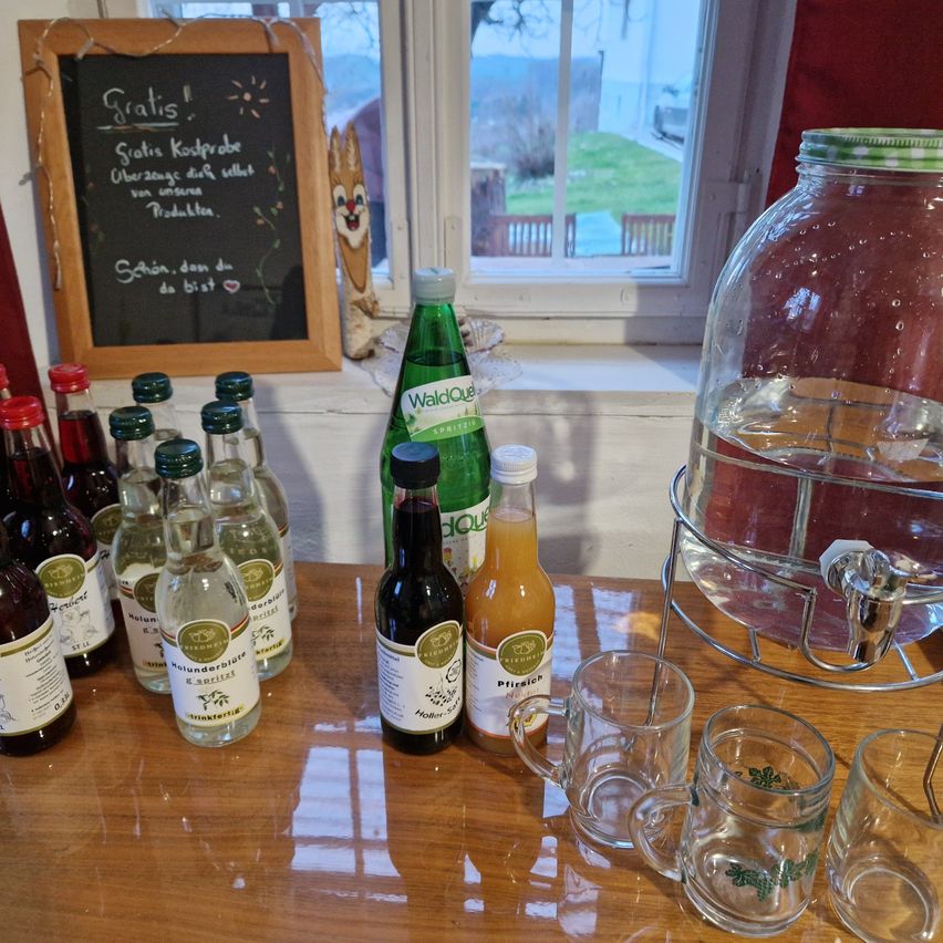 Ein Holztisch mit Flaschen, Gläsern und einem schwarzen Brett mit deutschem Text. Ein großer Krug mit grünem Deckel steht in der Nähe.