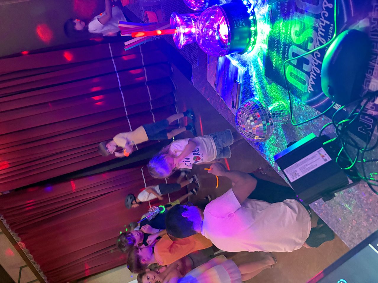 Eine Gruppe von Kindern und Erwachsenen befindet sich in einem dunklen Raum mit Disco-Lichtern und einer Discokugel. Die Kinder spielen mit Leuchtstäben und einige tragen Brillen.