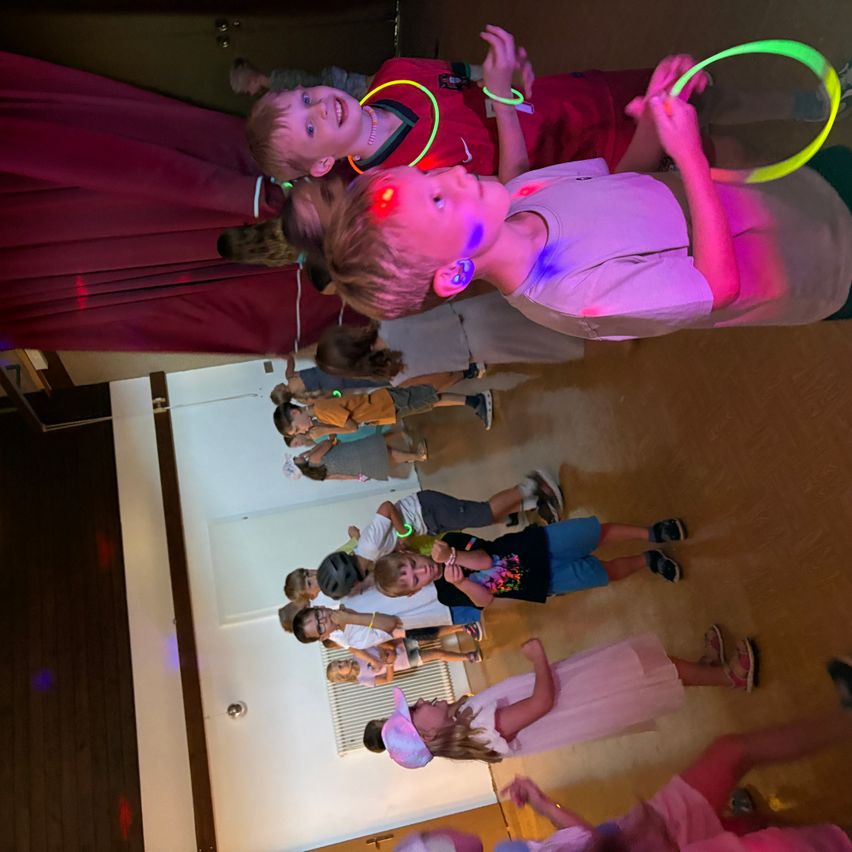 Eine Gruppe von Kindern spielt mit Leuchtstäben in einem Raum mit roter Beleuchtung. Einige stehen, andere sitzen auf dem Boden.