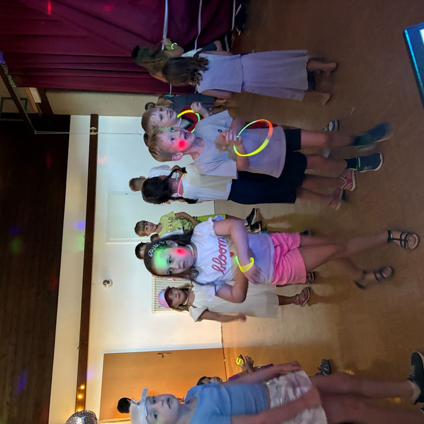 Eine Gruppe von Kindern mit Leuchtaccessoires und Gesichtsbemalung tanzt in einem Raum mit einer Discokugel und Discolichter.