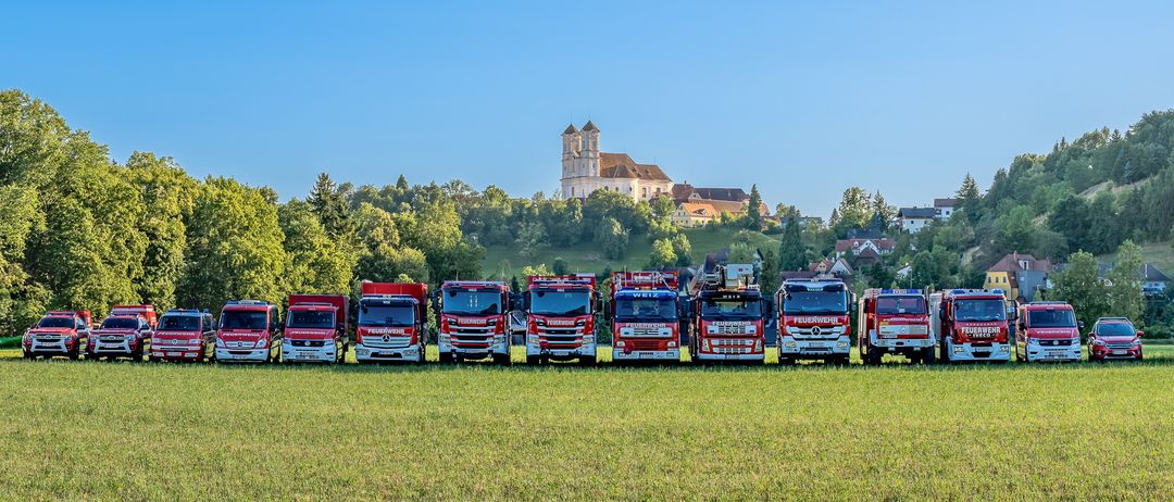 Eine Reihe roter Mercedes-Benz Feuerwehrfahrzeuge, die auf einer Wiese geparkt sind, mit einem Schloss auf einem Hügel im Hintergrund.