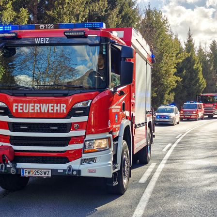 Ein roter Feuerwehrwagen mit 'FEUERWEHR' auf der Front ist auf der Straße geparkt. Dahinter fahren mehrere Einsatzfahrzeuge, einschließlich Polizeiautos. Bäume säumen die Straße auf beiden Seiten.