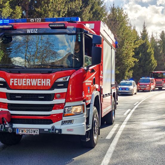 Ein roter Feuerwehrwagen mit 'FEUERWEHR' auf der Front ist auf der Straße geparkt. Dahinter fahren mehrere Einsatzfahrzeuge, einschließlich Polizeiautos. Bäume säumen die Straße auf beiden Seiten.