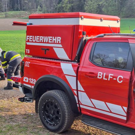 Ein roter Feuerwehrwagen steht auf einem Grasfeld, ein Feuerwehrmann repariert etwas am Wagen. Der Wagen hat eine große Lagerbox auf der Rückseite.