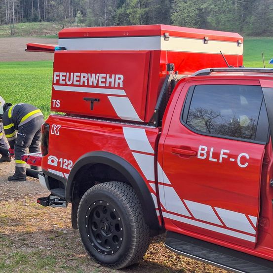Ein roter Feuerwehrwagen steht auf einem Grasfeld, ein Feuerwehrmann repariert etwas am Wagen. Der Wagen hat eine große Lagerbox auf der Rückseite.