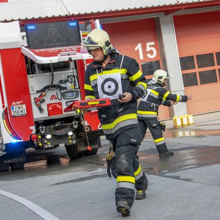 Zwei Feuerwehrleute gehen vor einem Feuerwehrauto. Einer hält ein Gerät, und der andere winkt. Das Auto hat ein Kennzeichen, das 356 WZ liest.