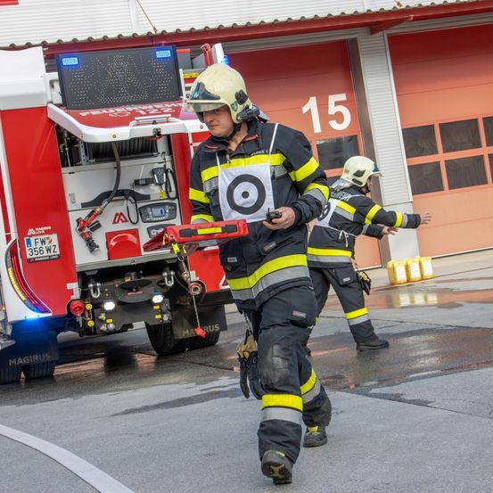 Zwei Feuerwehrleute gehen vor einem Feuerwehrauto. Einer hält ein Gerät, und der andere winkt. Das Auto hat ein Kennzeichen, das 356 WZ liest.