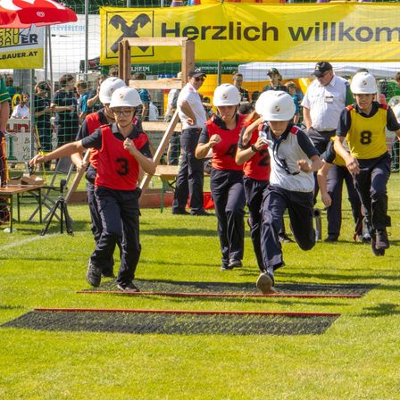 Eine Gruppe junger Personen läuft auf einem Rasenfeld. Sie tragen Helme und rote Westen. Über ihnen befindet sich ein Schild mit der Aufschrift 'Herzlich willkommen'.