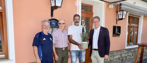 Vier Männer stehen vor einem rosa Gebäude. Ein Mann hält eine Topfpflanze und ein anderer trägt ein blaues Shirt mit Logo. Hinter ihnen ist eine Holztür und ein Schild zu sehen.