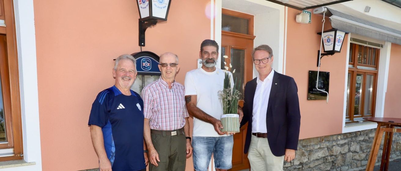Vier Männer stehen vor einem rosa Gebäude. Ein Mann hält eine Topfpflanze und ein anderer trägt ein blaues Shirt mit Logo. Hinter ihnen ist eine Holztür und ein Schild zu sehen.