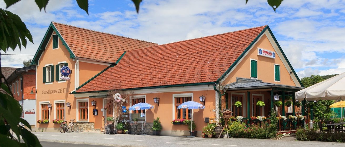 Ein kleines Hotel mit rotem Dach und grüner Einfassung. Es hat ein Schild über dem Eingang. Es hat viele Pflanzen und Blumen. Vorne stehen zwei blaue Regenschirme.