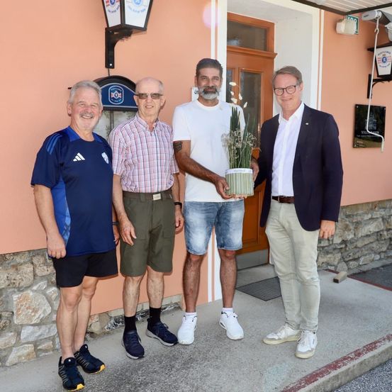 Vier Männer stehen vor einem rosa Gebäude mit einer Tür und lächeln. Einer hält eine Topfpflanze.