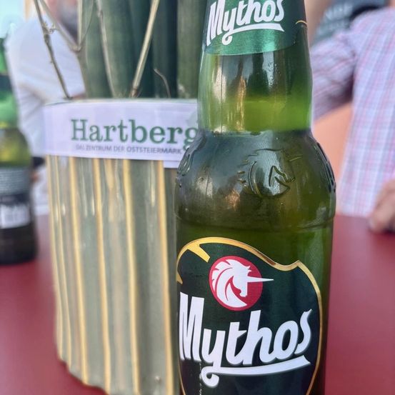 Eine Flasche Mythos Bier steht auf einem roten Tisch, mit einer Hartberg-Pflanze im Hintergrund. Die Flasche ist grün und trägt das Mythos-Logo. Eine weitere Flasche ist links teilweise sichtbar.