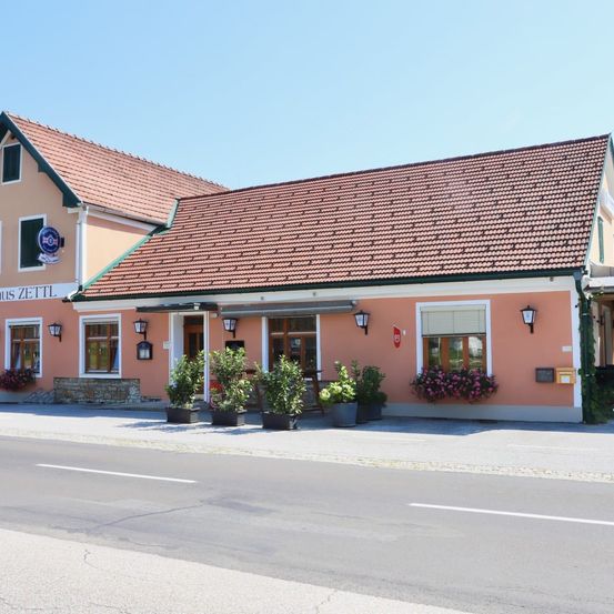 Ein rosafarbenes Gebäude mit rotem Dach und grünem Rand hat ein Schild mit der Aufschrift 'US ZETTL'. Das Gebäude hat mehrere Fenster und vor der Fassade gepflanzte Töpfe.