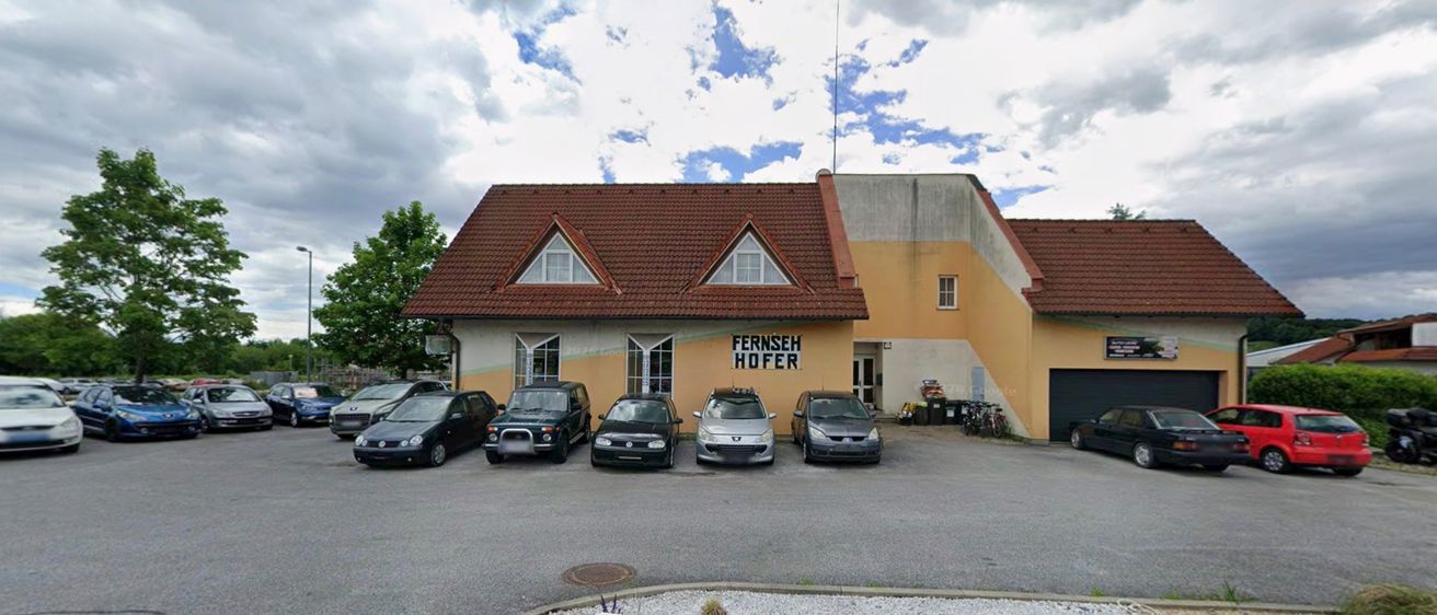 Ein gelbes Gebäude mit einem roten Dach und dem Schild 'Fernseh Hofer' hat mehrere Autos davor geparkt. Der Himmel ist bewölkt.