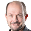 Ein lächelnder Mann mittleren Alters mit vollem Bart und Schnurrbart, der ein gemustertes schwarzes Hemd trägt. Er hat hellblaue Augen und kurzes, grau werdendes Haar.
