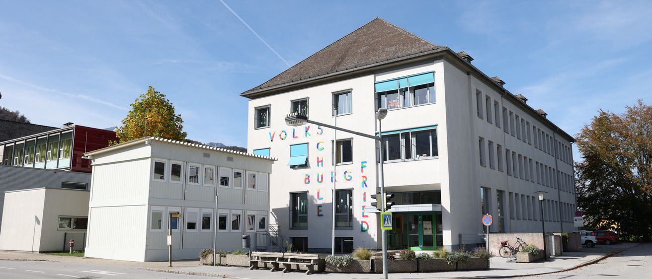 Ein großes weißes Gebäude mit braunem Dach hat große bunte Buchstaben, die 'Volkshochschule' auf seiner Seite buchstabieren. Es gibt mehrere Fenster am Gebäude. In der Nähe stehen Bänke und ein Fahrrad.
