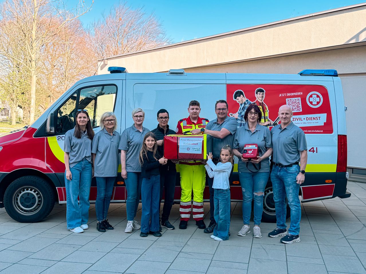 A group of people stands in front of an ambulance, holding a box. They are wearing uniforms and posing for a photo.