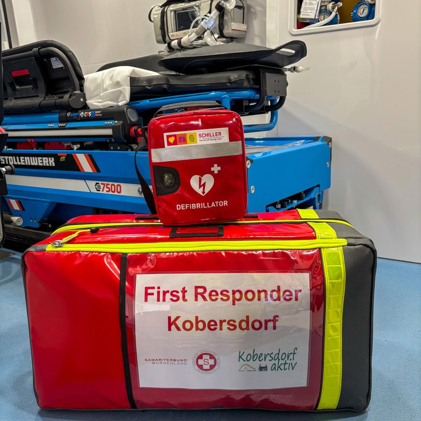 A red first responder bag from Kobersdorf sits on a blue stretcher. A defibrillator rests on top of the bag. A blue and white stretcher is in the background.