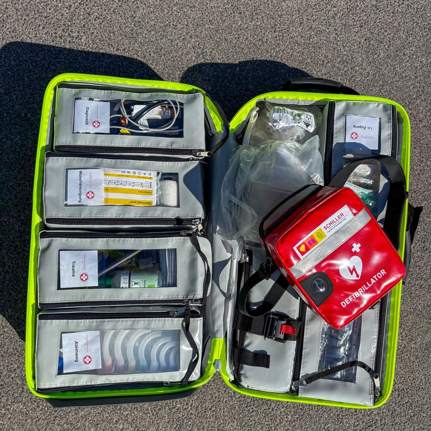 An open green and gray case with compartments and medical supplies, including a red defibrillator bag, is on the ground.
