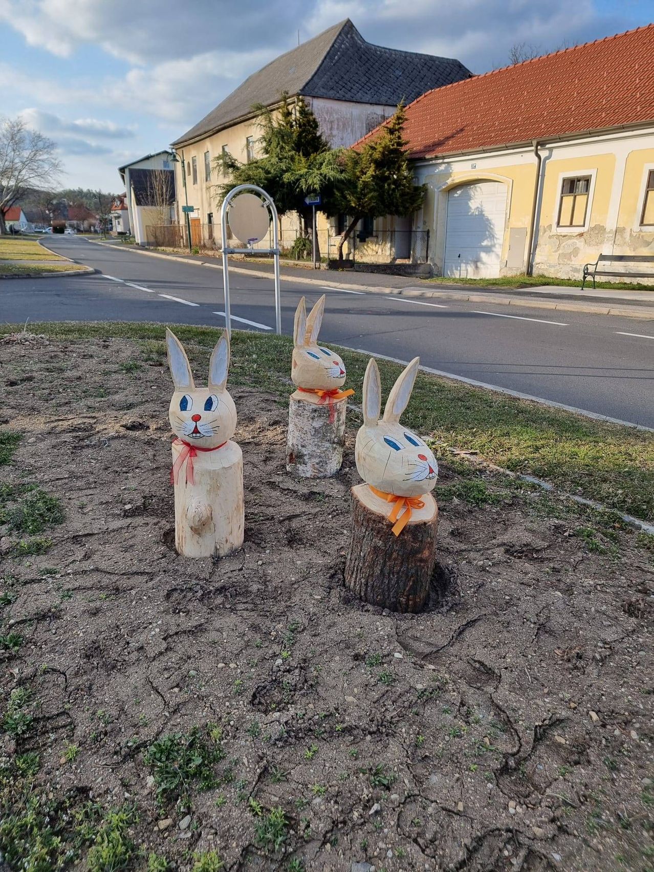 Drei geschnitzte Holzhasenfiguren stehen auf Baumstümpfen in einem Grasbereich neben einer Straße. Dahinter befindet sich ein Gebäude mit einem roten Dach und einer Bank auf dem Bürgersteig.
