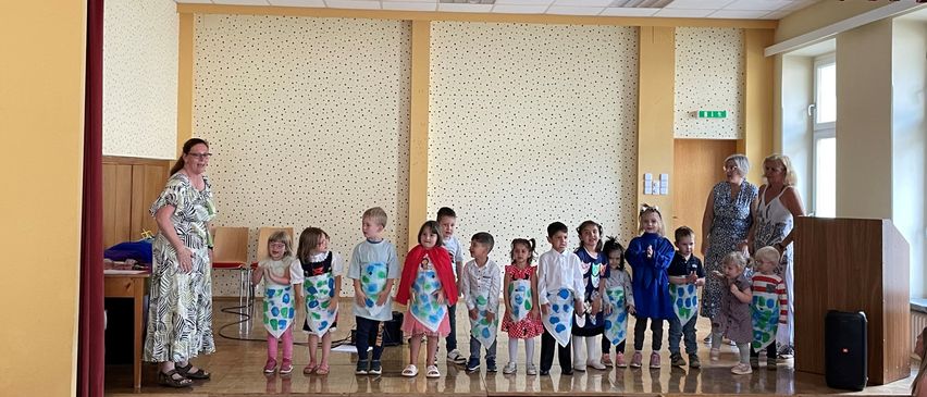 Eine Gruppe von Kindern steht auf der Bühne und trägt Schürzen, während eine Frau links steht. Hinter ihnen sind zwei Frauen anwesend, eine hält ein Baby. Der Raum hat einen Holzboden, Wände mit weißen Punkten und einen roten Vorhang oben.