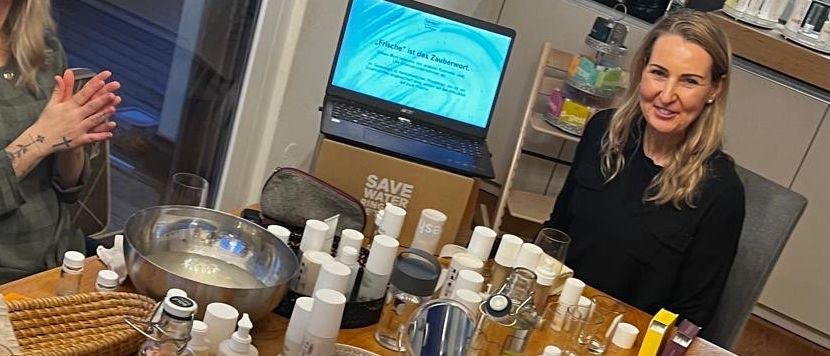 Vier Frauen sitzen um einen Holztisch herum mit verschiedenen Produkten, Flaschen und Gläsern. Ein Laptop und eine Schüssel befinden sich ebenfalls auf dem Tisch. Sie scheinen in einer Besprechung zu sein.