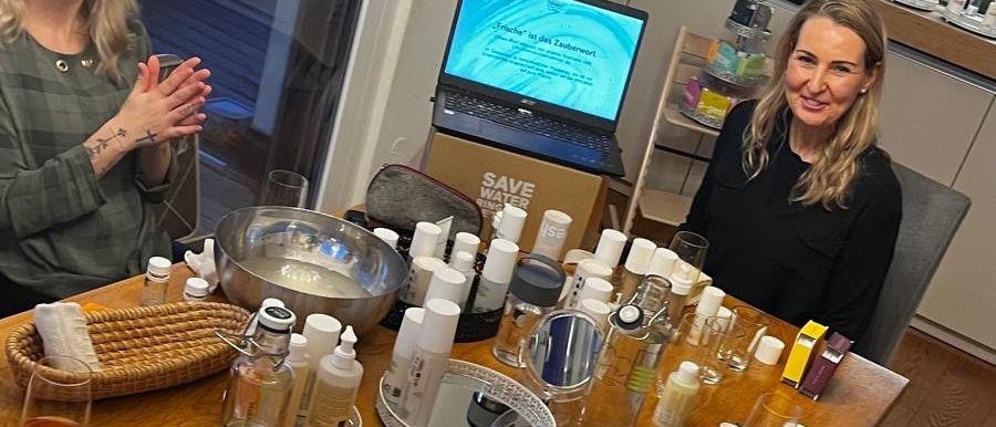 Drei Frauen sitzen um einen Tisch herum mit verschiedenen Flaschen und einem Laptop. Eine Frau lächelt und klatscht, eine andere schaut auf den Laptop-Bildschirm und die dritte konzentriert sich auf den Tisch.