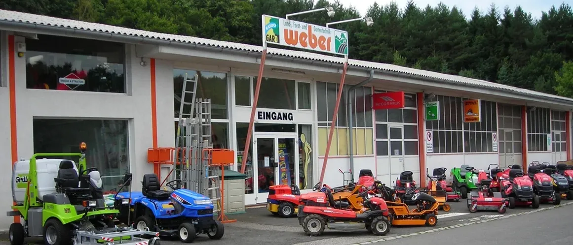 Das Bild zeigt ein Gebäude mit einem Schild, das 'Gartenfachmarkt Weber' liest. Draußen sind mehrere Rasenmäher geparkt, einige blau und einige rot. Im Hintergrund sind Bäume.