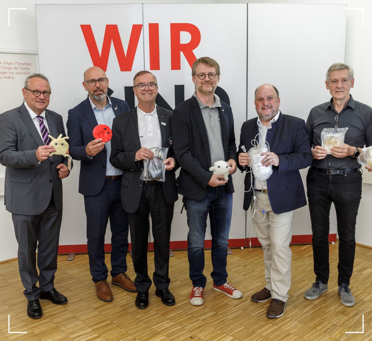 Fünf Männer in Anzügen und Turnschuhen stehen in einer Reihe und halten Kuscheltiere und Plastikbeutel, möglicherweise bei einer Pressekonferenz.