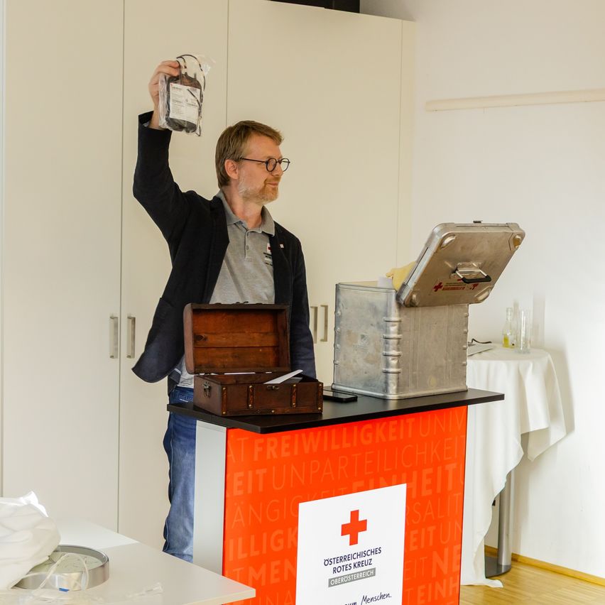 Ein Mann mit Brille hält eine Blutspendetüte in einem Raum mit weißer Tischdecke, Holzkiste und rotem Kreuz-Schild an der Wand.