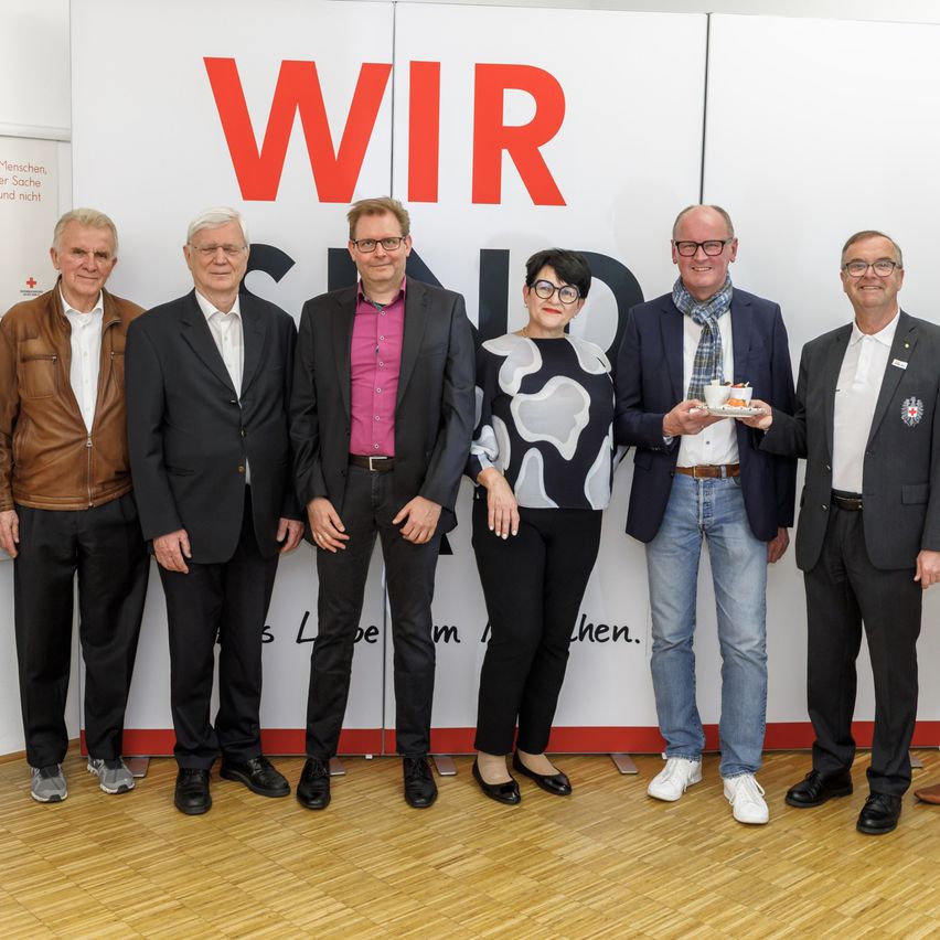 Sechs Männer und eine Frau stehen vor einer weißen Wand mit dem Text 'WIR' in roten Buchstaben. Sie sind in Anzügen und Schuhen gekleidet. Der Mann ganz rechts hält eine Tasse mit Untertasse.