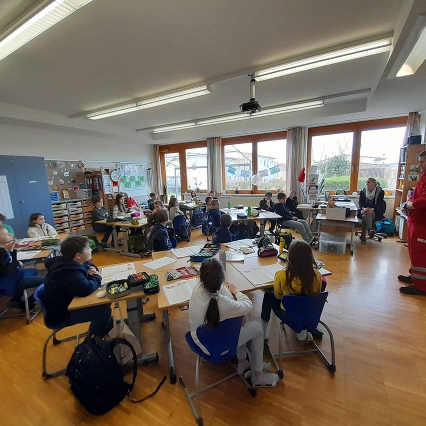 Ein Klassenzimmer mit Kindern an Tischen, einem Lehrer im Hintergrund und einem Feuerwehrmann in roter Kleidung.