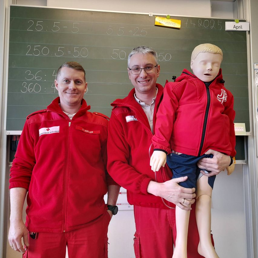 Zwei Männer in roten Jacken und ein Mann, der eine Kinderpuppe hält, stehen vor einer Tafel mit Zahlen und Gleichungen.