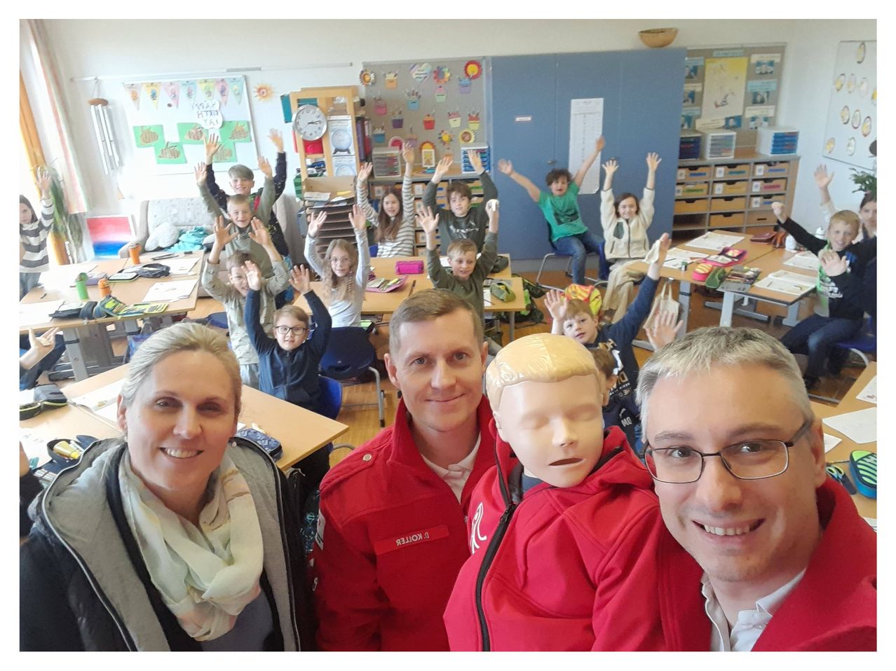 Drei Erwachsene und mehrere Kinder in einem Klassenzimmer. Die Kinder heben ihre Hände, und die Erwachsenen lächeln.