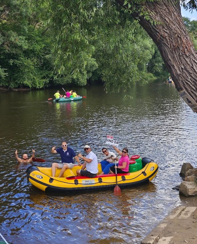 Eine Gruppe von Menschen befindet sich auf einem Schlauchboot im Fluss. Einige paddeln, während andere ein Schild halten. Ein Mann schwimmt in der Nähe. Ein weiteres Schlauchboot ist im Hintergrund zu sehen.