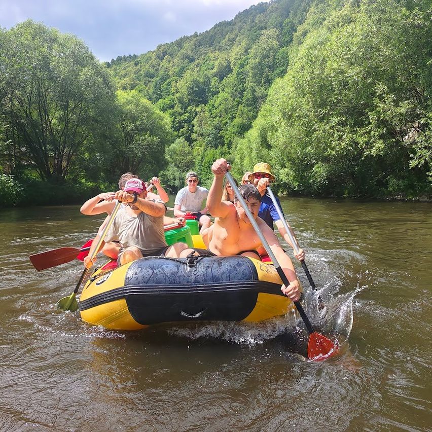 Eine Gruppe von Menschen fährt auf einem Fluss mit Schlauchbooten, trägt Hüte und hält Paddel. Bäume und Berge umgeben das Wasser.
