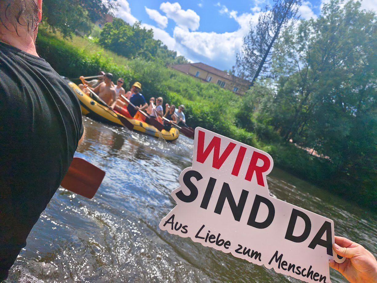 Eine Gruppe von Menschen treibt auf einem Fluss. Im Vordergrund steht ein Schild mit der Aufschrift 'Wir sind da'. Bäume und ein Haus sind im Hintergrund.