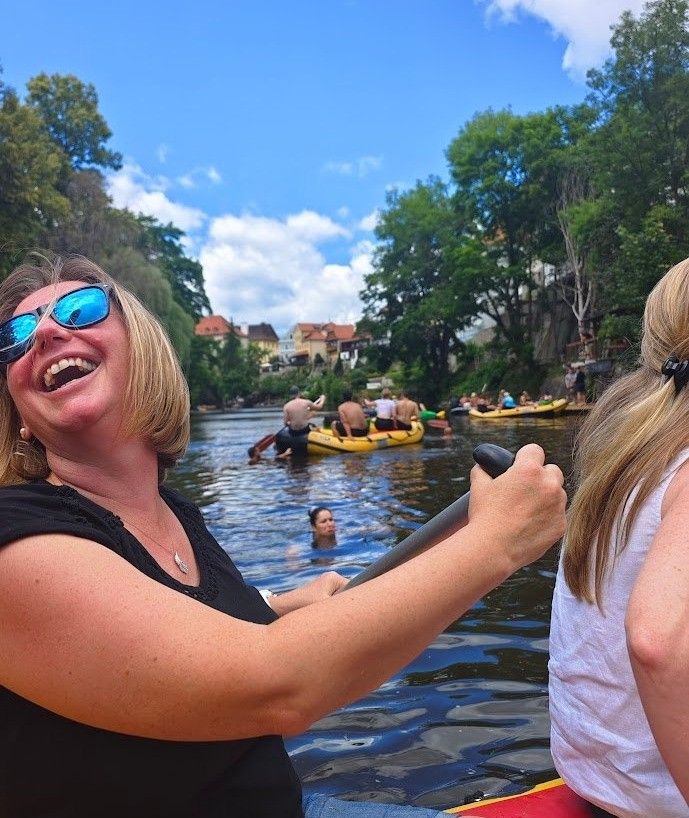 Eine Frau paddelt mit einem Floß auf einem Fluss, während andere raffahren und schwimmen. Bäume und Gebäude säumen die Ufer unter einem blauen Himmel mit Wolken.