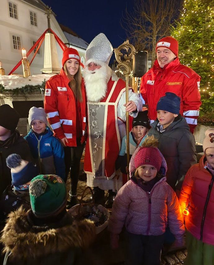 Der Weihnachtsmann steht vor einer Gruppe von Kindern und zwei Erwachsenen in roten Jacken, mit einem Weihnachtsbaum und einer Kirche im Hintergrund.