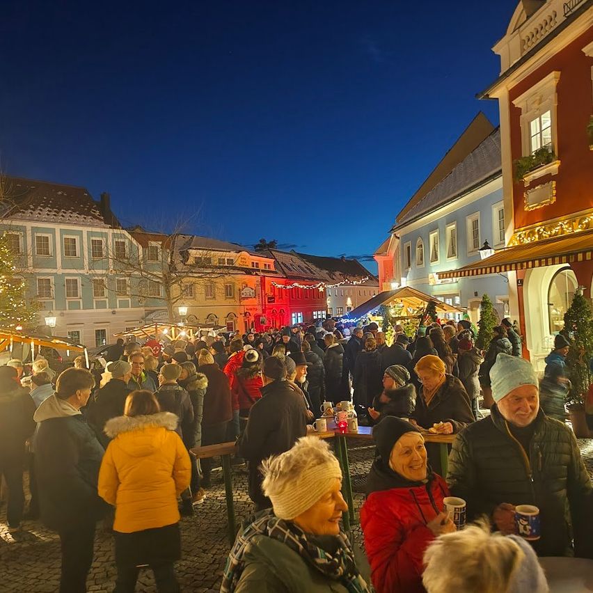 Bei Nacht ist ein überfüllter Straßenmarkt mit Menschen belebt, einige in Winterkleidung, andere mit Mützen, genießen die festliche Atmosphäre. Einige Menschen sitzen an einem Tisch, und ein großer Weihnachtsbaum ist vorhanden.