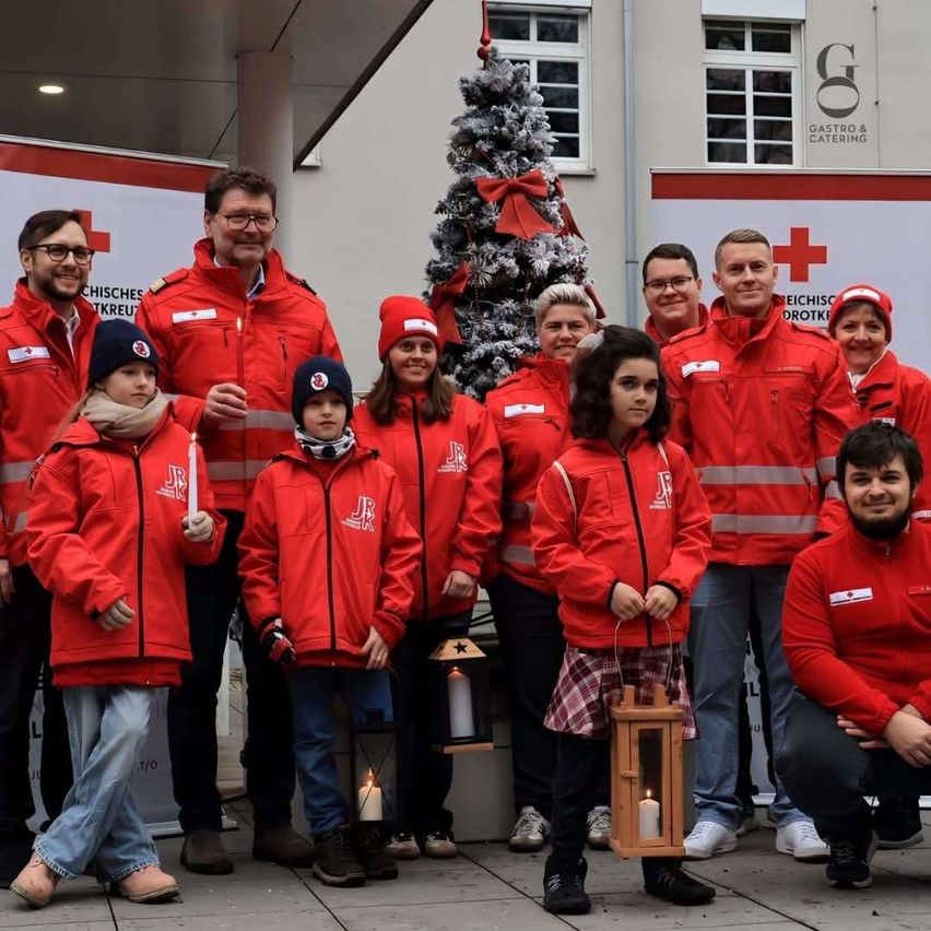 Eine Gruppe von Menschen in roten Jacken posiert für ein Foto vor einem Weihnachtsbaum. Sie halten Kerzen und lächeln. Dahinter befindet sich ein Banner mit einem Logo und Text.