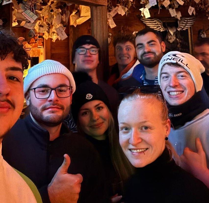 Eine Gruppe von Menschen, darunter Männer und Frauen, lächelt und posiert für ein Selfie in einem Raum. Sie tragen Winterkleidung.