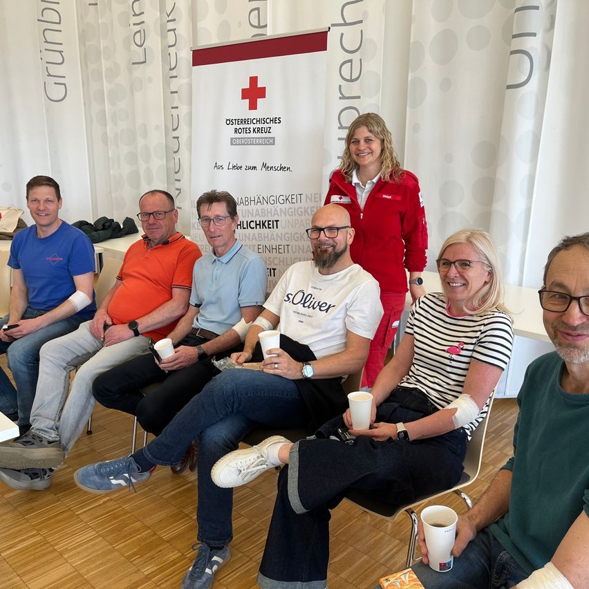 Gruppe von lächelnden Menschen, einige mit Verbänden, sitzen auf Stühlen, einer steht in einem roten Jackett. Im Hintergrund ein Banner mit der Aufschrift 'Österreichisches Rotes Kreuz'. Holzboden.