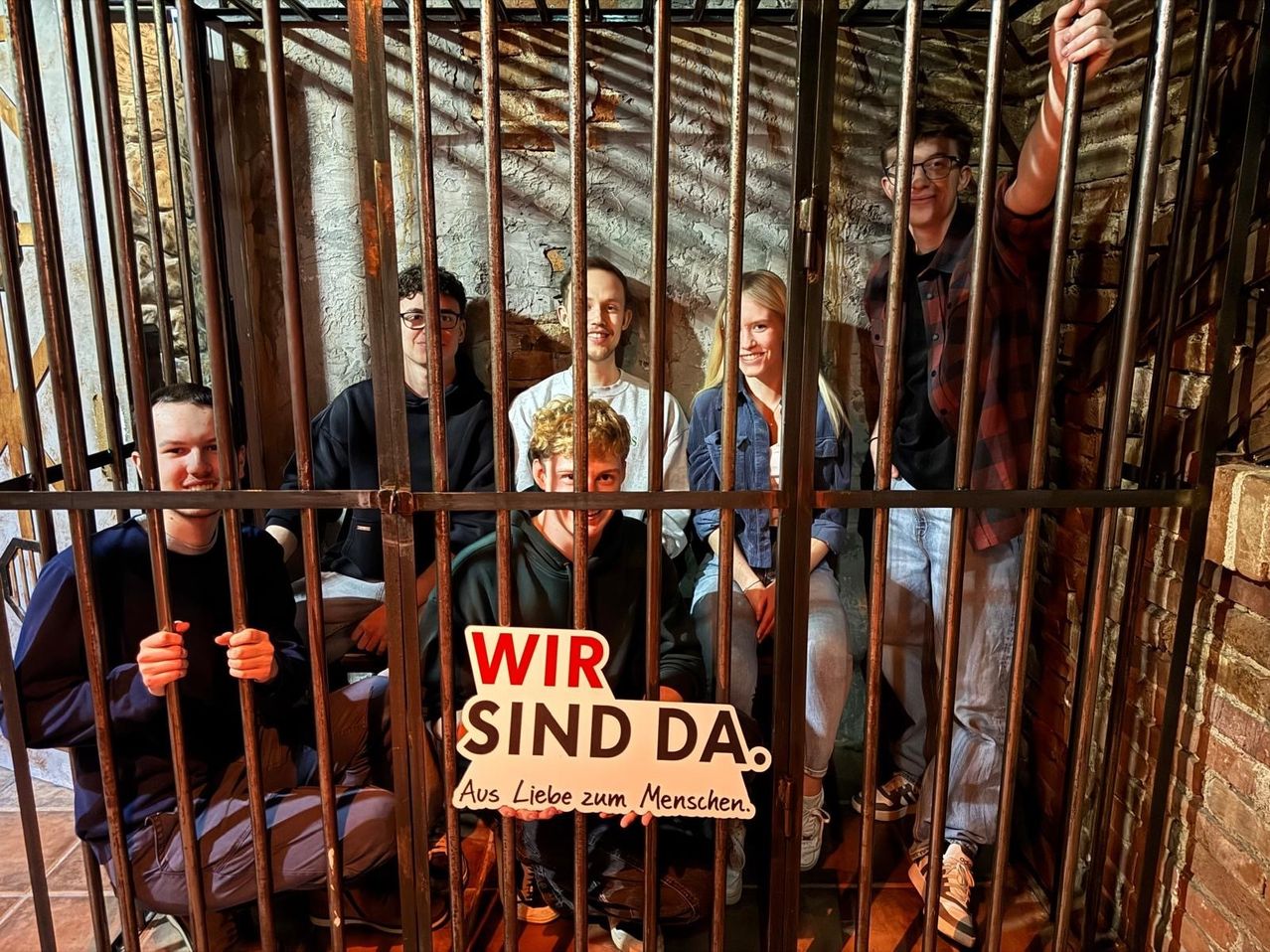 A group of people are inside a mock prison cell, smiling and posing for a photo. A sign in front reads 'Wir sind da'.